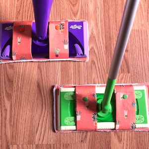 May include: Two Swiffer mop heads with a floral pattern. The mop heads are orange with green plants and the text "Swiffer" printed on the top. One mop head is attached to a purple handle and the other is attached to a silver handle.