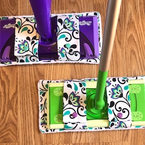 May include: Two Swiffer mop pads with a floral design in black, white, green, and purple. The pads are attached to the mop handles, which are purple and green.