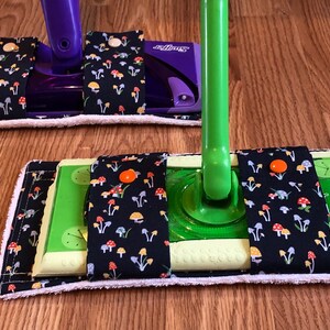 May include: Two Swiffer Sweeper mop pads with a black fabric cover with a mushroom print. The fabric has orange, yellow, and blue mushrooms on a black background. The pads are attached to the mop handles with velcro.