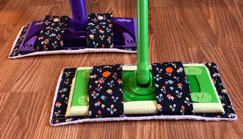 May include: Two Swiffer Sweeper mop pads with a black fabric cover with a mushroom print. The fabric has orange, yellow, and blue mushrooms on a black background. The pads are attached to the mop handles with velcro.