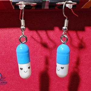 May include: A pair of novelty earrings featuring blue and white pill-shaped charms with smiling faces and heart accents. The earrings hang from silver-colored fish hook ear wires, perfect for adding a touch of whimsy to any outfit.