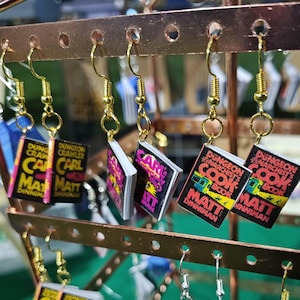 May include: Dangle earrings featuring miniature book charms with black covers and yellow text. The charms display the titles Dungeon Crawler Carl and Dungeon Anarchist's Cook Book by Matt Dinniman. Each charm hangs from a gold-toned metal fishhook wire.