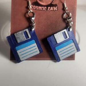 May include: A pair of earrings featuring a retro floppy disk design. The earrings are blue with silver and light blue accents, and hang from silver-colored hooks. The background includes a brown card with the text "COSMIC CAFE".