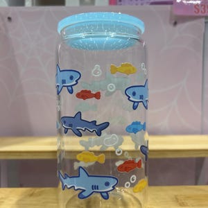 May include: Clear glass water bottle with a light blue lid. The bottle is decorated with cartoon sharks, yellow, red, and blue fish, and bubbles. The bottle is cylindrical and sits on a wooden shelf.