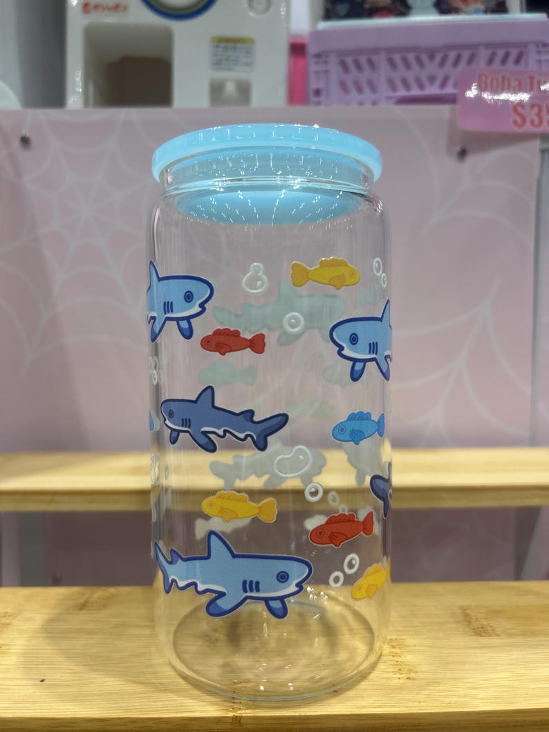 May include: Clear glass water bottle with a light blue lid. The bottle is decorated with cartoon sharks, yellow, red, and blue fish, and bubbles. The bottle is cylindrical and sits on a wooden shelf.