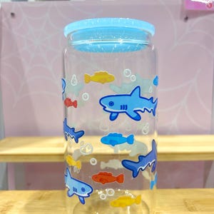 May include: A clear glass drinking jar with a light blue lid. The jar is decorated with cartoon sharks and fish in shades of blue, yellow, and red. The jar is cylindrical and appears to be empty.