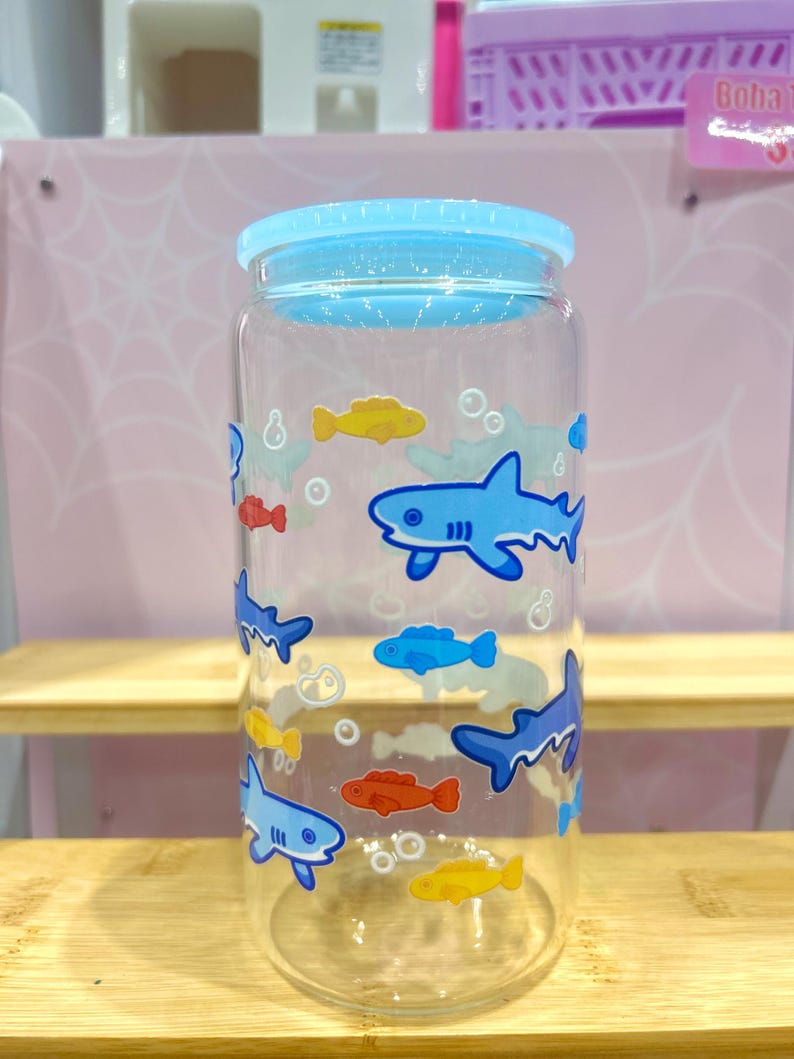 May include: A clear glass drinking jar with a light blue lid. The jar is decorated with cartoon sharks and fish in shades of blue, yellow, and red. The jar is cylindrical and appears to be empty.