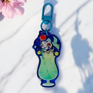 Puede incluir: Un llavero acrílico azul con un diseño de cóctel de dibujos animados. El llavero tiene un cierre azul y una forma de vaso de cóctel con una bebida verde, una cereza y un personaje de dibujos animados. El fondo es blanco con flores rosas.