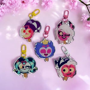 May include: Five colorful cartoon character keychains with a popsicle design. Each keychain features a different character with a unique expression and color scheme. The keychains are made of acrylic and have a silver metal ring.