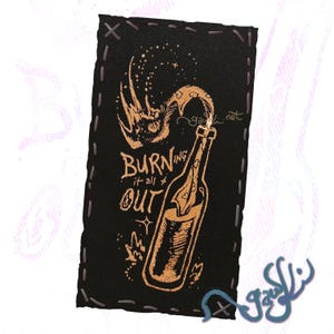 May include: Black rectangular patch with a gold illustration of a bottle with flames and the text "BURNING it all OUT". The patch has a stitched border and a stylized design.