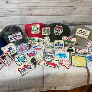 May include: A collection of embroidered patches featuring various designs and phrases, including "Farm Life", "Naughty but Too Cute to Cart", "Aloha Beaches", "Super Mom Super Wife Super Tired", "Pumpkin Spice", and "Chaos Coordinator". The patches are displayed on a light brown surface with several baseball caps.