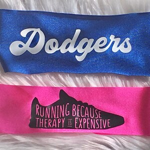 K&ouml;nnte beinhalten: Zwei Stirnb&auml;nder, eines blau mit dem Wort "Dodgers" in wei&szlig;en Buchstaben und eines pink mit einer schwarzen Silhouette eines Laufschuhs und dem Text "Running because therapy is expensive."