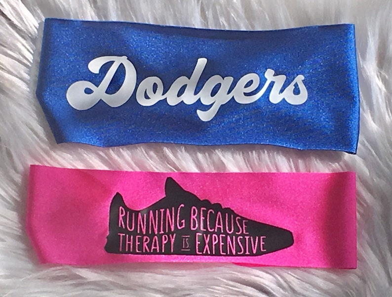 K&ouml;nnte beinhalten: Zwei Stirnb&auml;nder, eines blau mit dem Wort "Dodgers" in wei&szlig;en Buchstaben und eines pink mit einer schwarzen Silhouette eines Laufschuhs und dem Text "Running because therapy is expensive."