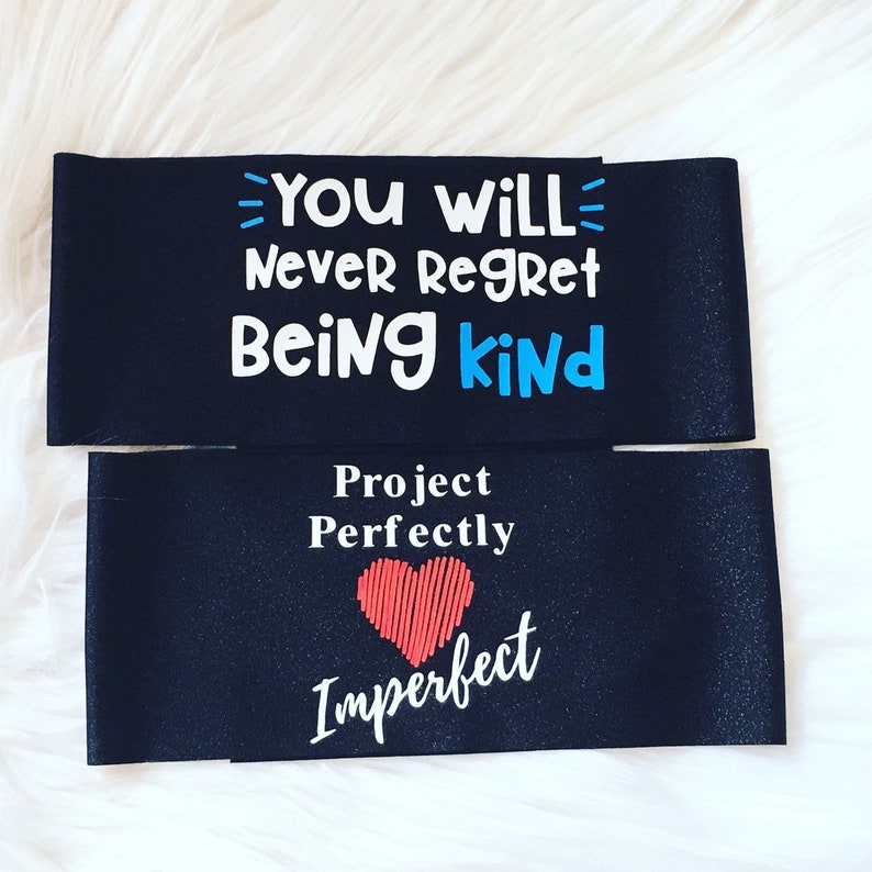 K&ouml;nnte beinhalten: Zwei schwarze Stoff-Getr&auml;nkedosenk&uuml;hler mit wei&szlig;em und blauem Text. Der obere K&uuml;hler sagt "You will never regret being kind". Der untere K&uuml;hler sagt "Project Perfectly Imperfect" mit einer roten Herzgrafik.