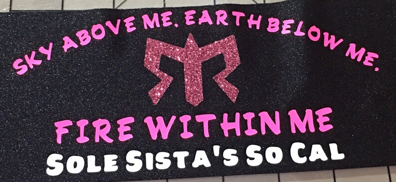 K&ouml;nnte beinhalten: Schwarzes Stoffarmband mit pinkfarbenem Glitzer-Schriftzug "SKY ABOVE ME, EARTH BELOW ME. FIRE WITHIN ME SOLE SISTA'S SO CAL". Das Armband hat ein pinkfarbenes Glitzer-Logo in der Mitte.