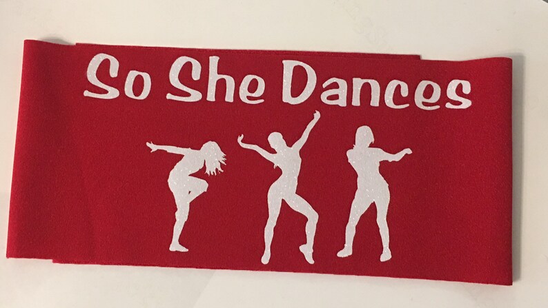 K&ouml;nnte beinhalten: Roter Stoffbanner mit wei&szlig;em Text "So She Dances" und drei wei&szlig;en Silhouetten von T&auml;nzern.