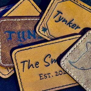 May include: Several rectangular leather patches in shades of brown are displayed. Some patches have text such as "Tynker" and "The Sm", while another features a ghost illustration and the words "Spooky Szn". The patches are stitched with white thread.