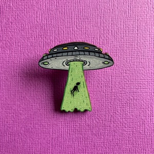 Puede incluir: Un pin de estilo caricatura que representa un OVNI gris y negro con luces amarillas. Un rayo verde emana del OVNI, que contiene una pequeña figura de alienígena negro. El pin está sobre un fondo morado texturizado.