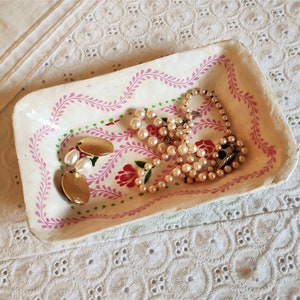 May include: A white ceramic trinket dish with pink and green floral designs. The dish contains a pearl necklace and a pair of gold earrings.