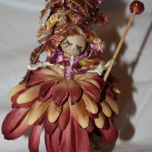 May include: A handmade doll with a burgundy flower skirt, brown and gold hair, and a painted face. The doll is holding a stick with a red glitter ball on top.