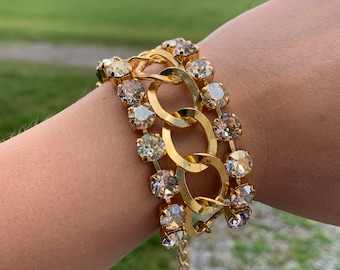 May include: A gold chain bracelet with clear and champagne-colored crystals. The bracelet is made of metal and has a clasp closure.