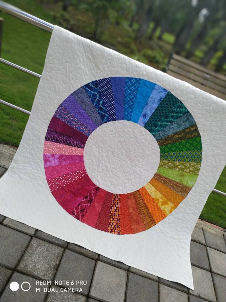 Op de afbeelding: Een wit quilt met een kleurrijk cirkelontwerp. De cirkel is gemaakt van verschillende stofresten in tinten paars, blauw, groen, geel, oranje en rood. De cirkel is gecentreerd op het quilt en is omgeven door een witte achtergrond.