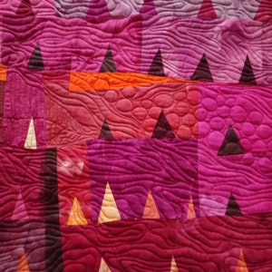 Op de afbeelding: Een quilt met een geometrisch patroon van driehoeken en vierkanten in tinten roze, rood, oranje en bruin. De quilt is gestikt met een golvend patroon.