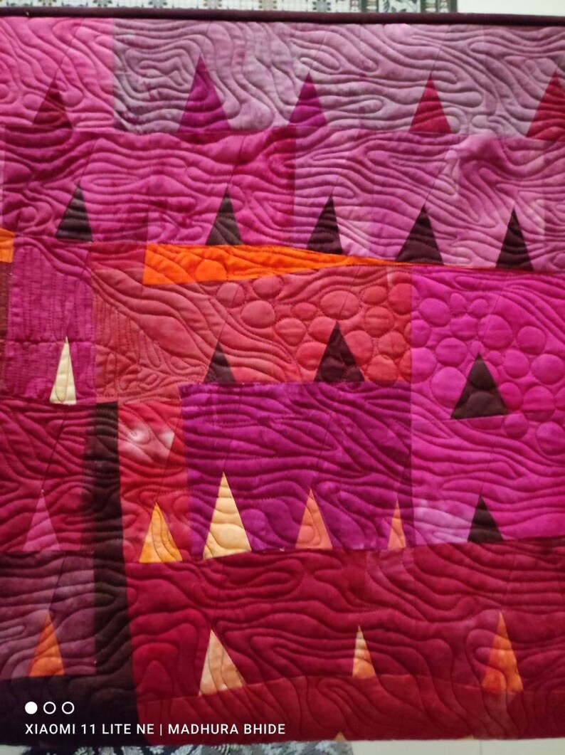 Op de afbeelding: Een quilt met een geometrisch patroon van driehoeken en vierkanten in tinten roze, rood, oranje en bruin. De quilt is gestikt met een golvend patroon.