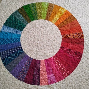 Op de afbeelding: Een wit quilt met een kleurrijk regenboogcirkelontwerp. De cirkel is gemaakt van verschillende stofresten die in een cirkelvormig patroon aan elkaar zijn genaaid. Het quilt is gestikt met een klein, herhalend patroon.
