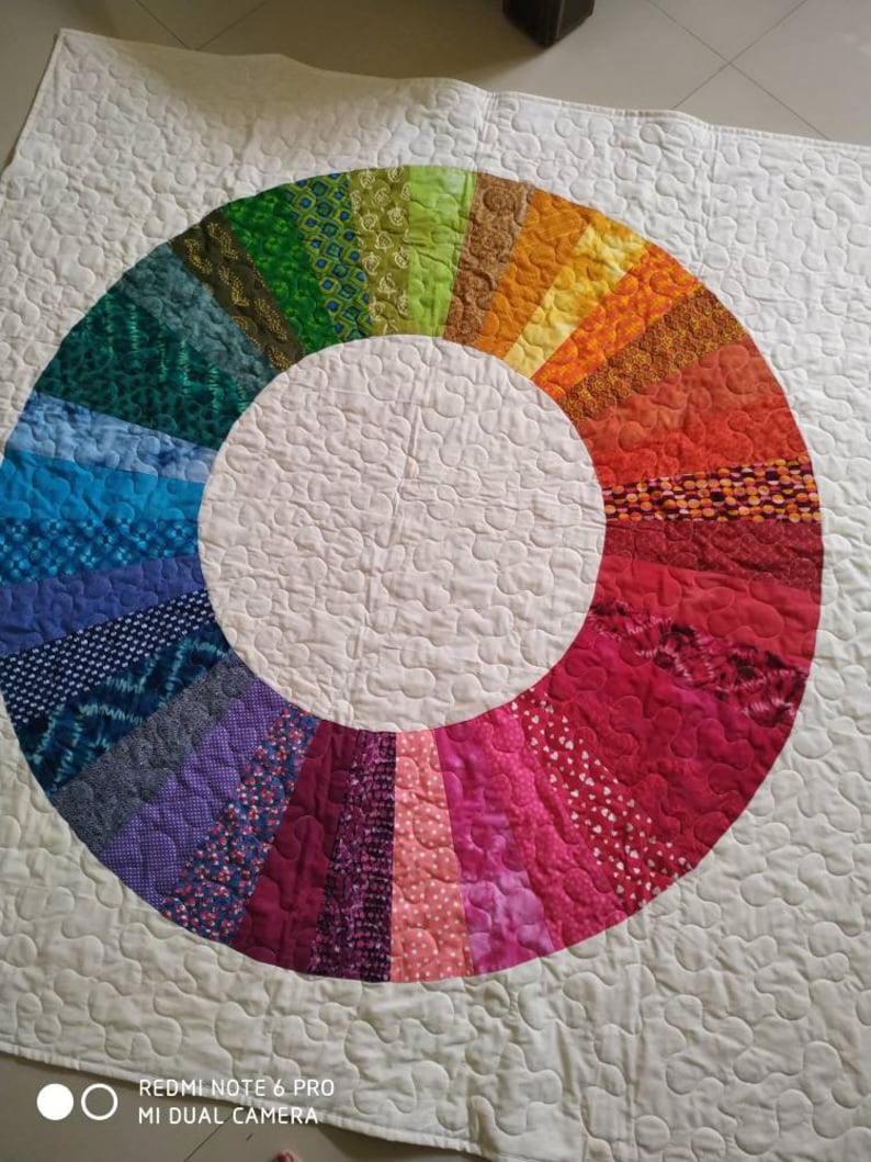 Op de afbeelding: Een wit quilt met een kleurrijk regenboogcirkelontwerp. De cirkel is gemaakt van verschillende stofresten die in een cirkelvormig patroon aan elkaar zijn genaaid. Het quilt is gestikt met een klein, herhalend patroon.