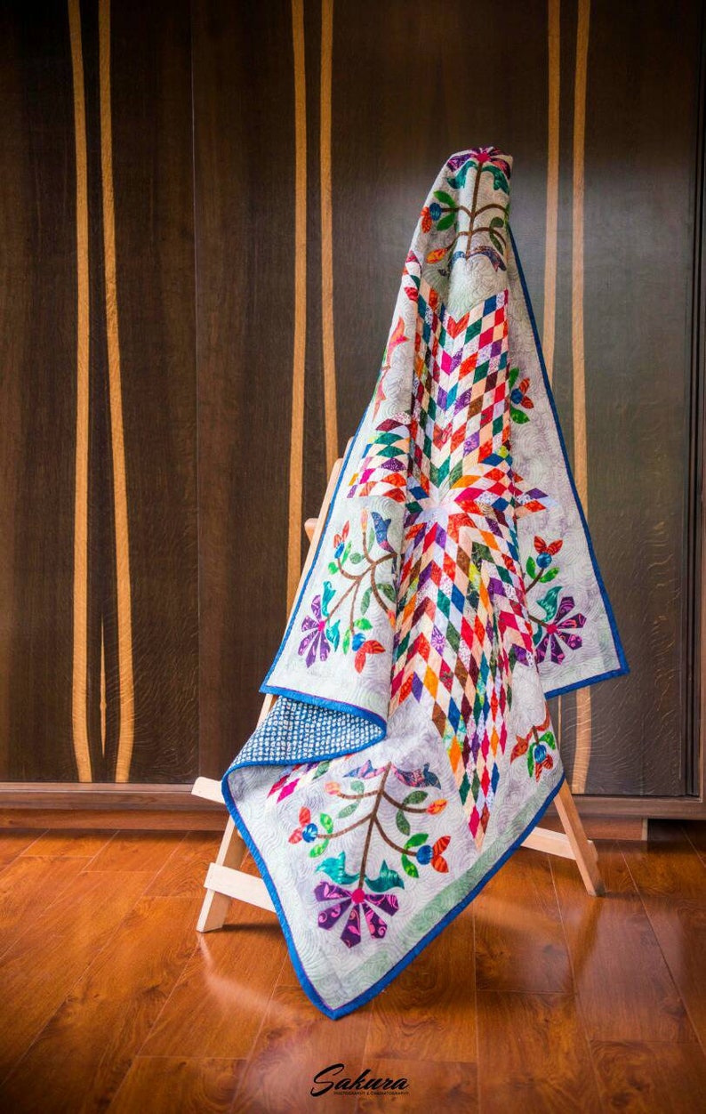 May include: A quilt featuring a central star pattern composed of multicolored diamond shapes in shades of red, orange, yellow, green, blue, and purple. The quilt has a light gray background with floral vine motifs and a solid blue border trim.