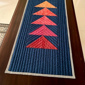 May include: A blue quilted table runner with a white border. The runner features five colorful triangles in shades of orange, red, and pink.
