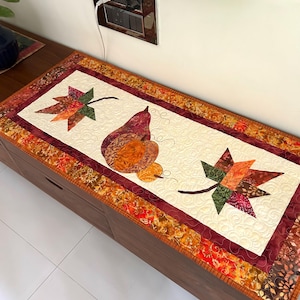 Puede incluir: Camino de mesa acolchado rectangular con diseño de pera y hojas. El centro es crema con una pera y dos hojas en tonos naranja, verde y marrón. El borde es una mezcla de telas estampadas en colores otoñales.