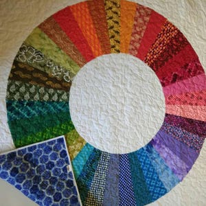 Op de afbeelding: Een wit quilt met een kleurrijk cirkelvormig ontwerp gemaakt van stofresten. Het ontwerp is een regenboog van kleuren, waarbij elke kleur wordt weergegeven door een ander stoffen patroon. Het quilt is doormidden gevouwen, waardoor een blauwe stoffen achterkant met een honingraatpatroon zichtbaar wordt.