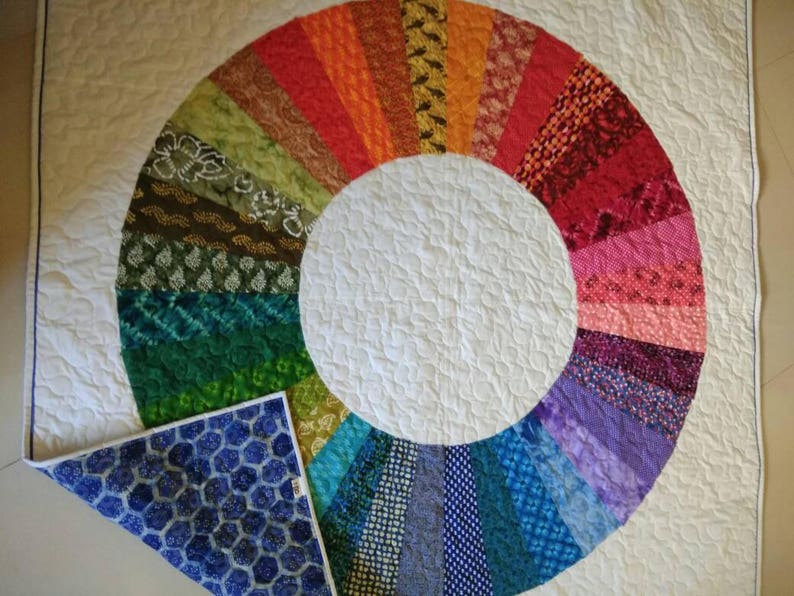 Op de afbeelding: Een wit quilt met een kleurrijk cirkelvormig ontwerp gemaakt van stofresten. Het ontwerp is een regenboog van kleuren, waarbij elke kleur wordt weergegeven door een ander stoffen patroon. Het quilt is doormidden gevouwen, waardoor een blauwe stoffen achterkant met een honingraatpatroon zichtbaar wordt.
