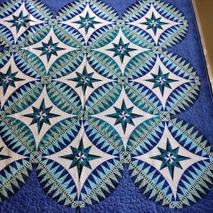 May include: A blue and white quilt with a geometric pattern of stars and circles. The quilt is made of different shades of blue and white fabric and has a quilted texture.