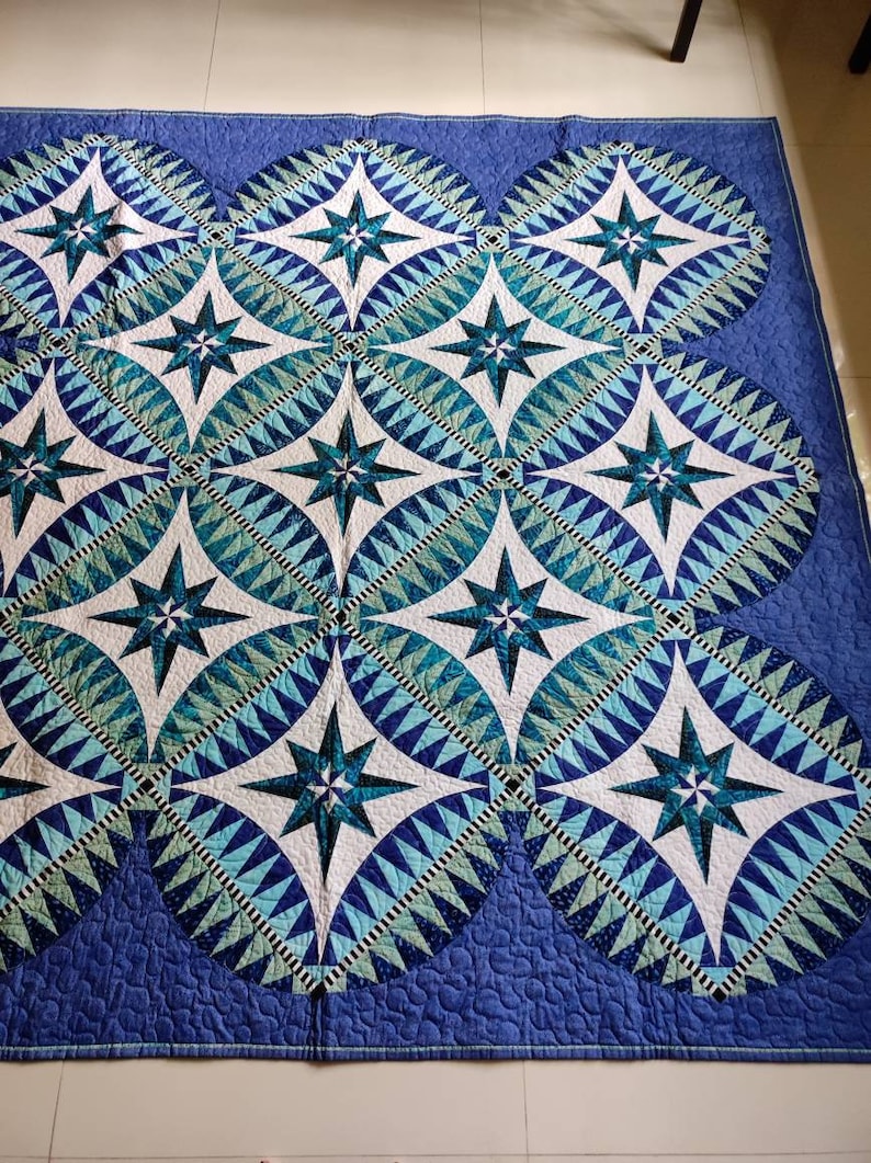 May include: A blue and white quilt with a geometric pattern of stars and circles. The quilt is made of different shades of blue and white fabric and has a quilted texture.