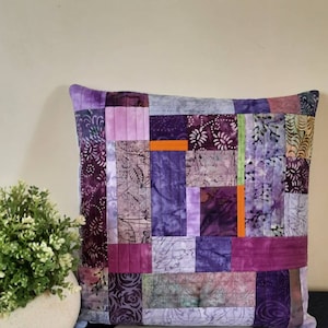 May include: Purple and lavender patchwork cushion with orange accents. The cushion is made of various fabrics with different patterns and textures.
