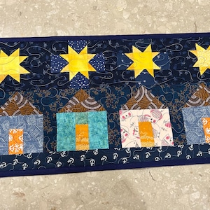 Könnte beinhalten: Quilt-Tischläufer mit Sternenhimmel-Design, gelben Sternen und vier Häusern. Die Häuser bestehen aus blauen, türkisfarbenen und gemusterten Stoffen mit orangefarbenen Türen, vor einem dunkelblauen Hintergrund.