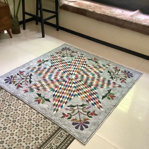 May include: A square quilt featuring a large central star composed of multicolored diamond-shaped fabric pieces. The star is set against a light gray background with intricate swirling quilting patterns and four floral motifs in the corners.