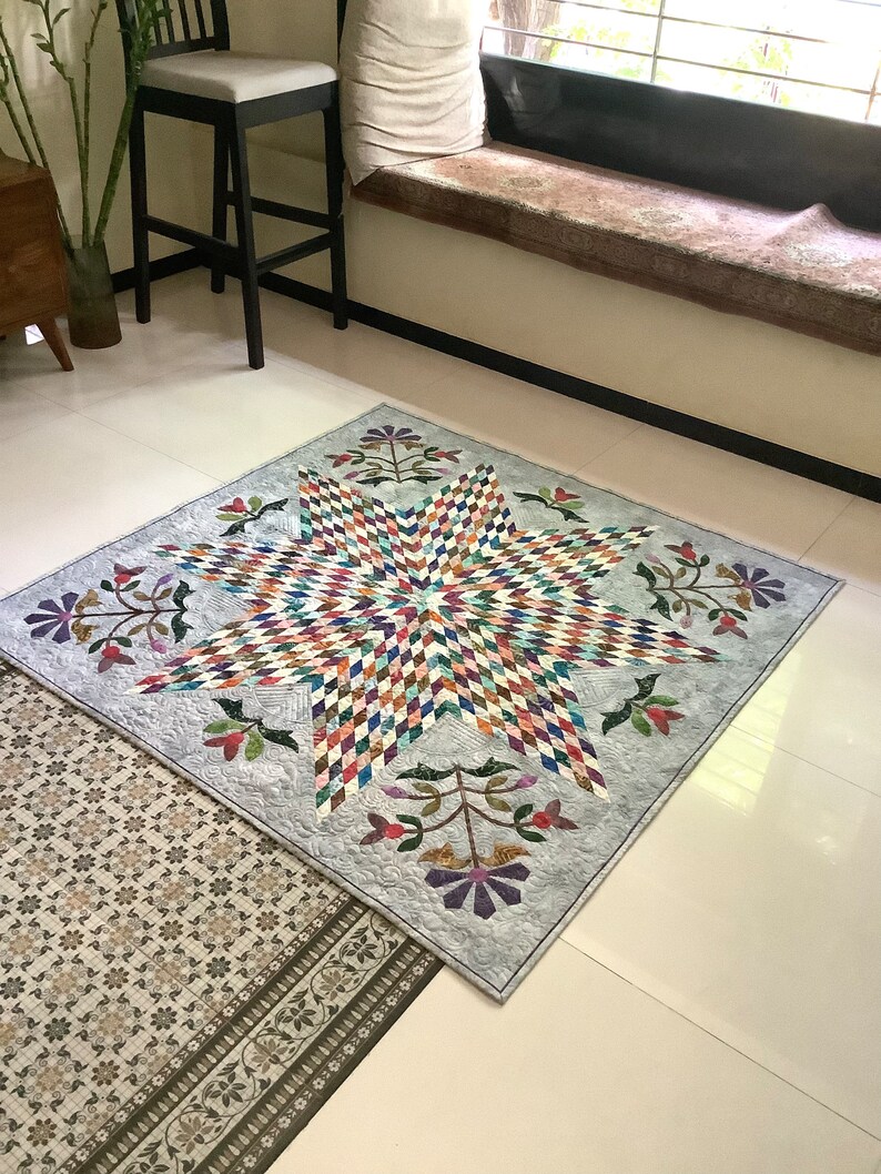 May include: A square quilt featuring a large central star composed of multicolored diamond-shaped fabric pieces. The star is set against a light gray background with intricate swirling quilting patterns and four floral motifs in the corners.