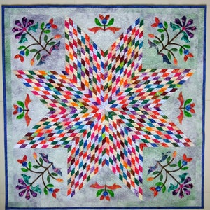 May include: A square quilt featuring a large central eight-pointed star composed of small multicolored diamond-shaped fabric pieces. The background is a light gray fabric with swirling quilted patterns and floral motifs in the corners.