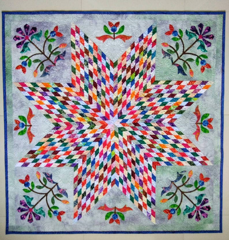 May include: A square quilt featuring a large central eight-pointed star composed of small multicolored diamond-shaped fabric pieces. The background is a light gray fabric with swirling quilted patterns and floral motifs in the corners.