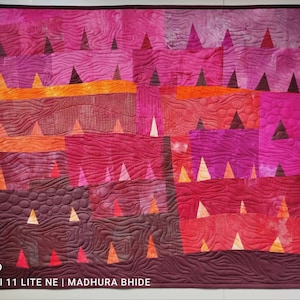 Op de afbeelding: Een quilt met een patchwork-ontwerp met verschillende tinten roze, rood en oranje. De quilt is gemaakt van driehoeken en vierkanten, en de stof heeft een textuur, gestikte look. De quilt is onderaan rechts gesigneerd met "Madhura Bhide".