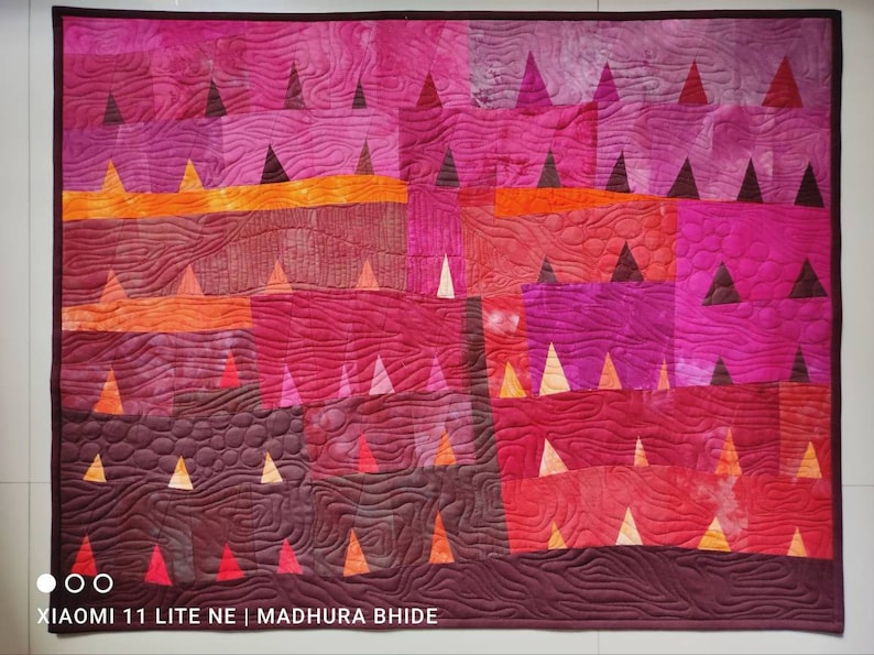 Op de afbeelding: Een quilt met een patchwork-ontwerp met verschillende tinten roze, rood en oranje. De quilt is gemaakt van driehoeken en vierkanten, en de stof heeft een textuur, gestikte look. De quilt is onderaan rechts gesigneerd met "Madhura Bhide".