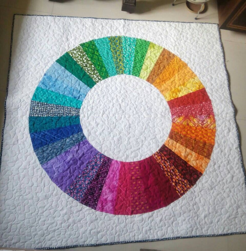 Op de afbeelding: Een wit quilt met een kleurrijk regenboogcirkelontwerp. De cirkel is gemaakt van verschillende stofvierkanten in tinten blauw, groen, geel, oranje, rood en paars.