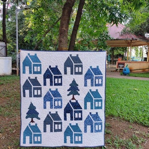 Puede incluir: Un edredón hecho a mano con un patrón repetido de casas y árboles azules sobre un fondo blanco. Las casas varían en tonos de azul, creando un diseño encantador. El edredón se exhibe al aire libre, mostrando su potencial como artículo decorativo para el hogar.