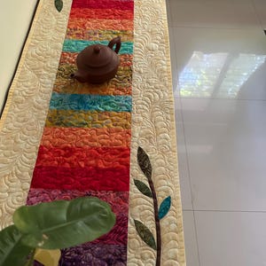 May include: A colorful quilted table runner with a cream background and a brown teapot on top. The runner has a pattern of leaves on the sides and a repeating pattern of swirls on the cream background.