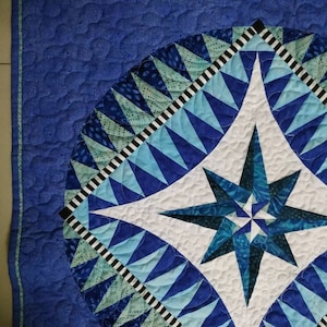 May include: A blue and white quilt with a starburst pattern in the center. The quilt is made of various shades of blue and white fabric and has a black and white border.