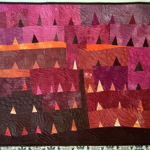Op de afbeelding: Een quilt met een geometrisch patroon van driehoeken in tinten bruin, oranje en roze. De quilt is gemaakt van vierkante stoffen die aan elkaar zijn genaaid om een patchwork-ontwerp te cre&euml;ren.